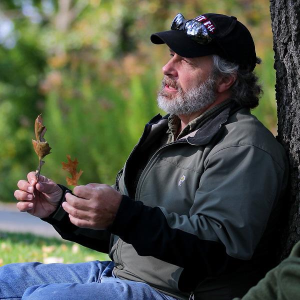 Tony Gulig sits with his back against a tree holding a leaf in each hand.