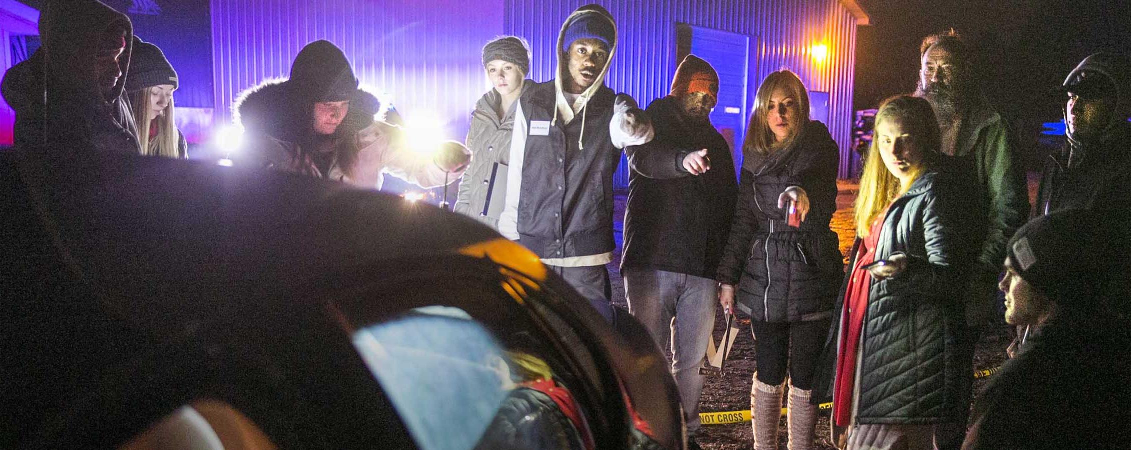 Students stand outside in the dark by a car during a crime scene simulation.