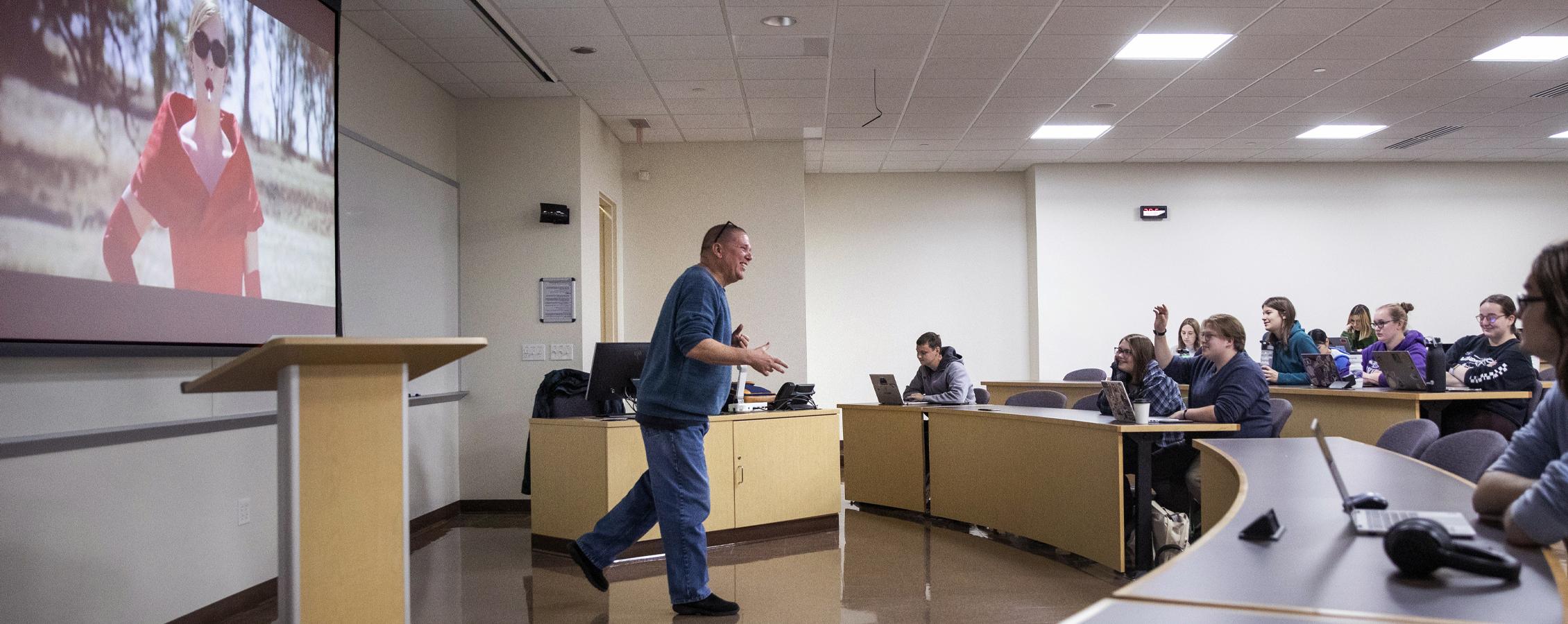 A faculty member speaks at the front of class while a film plays behind him.