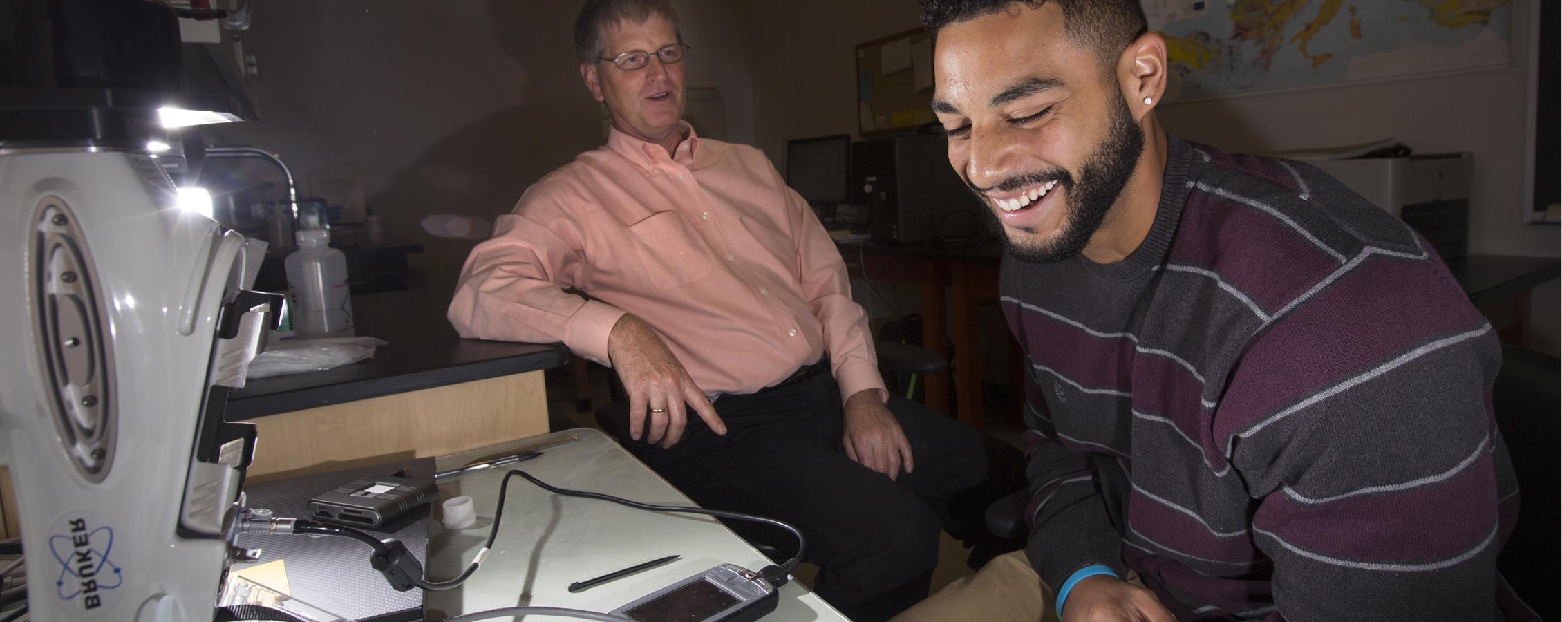 A student is indoors with his professor, using technology to test soil samples.