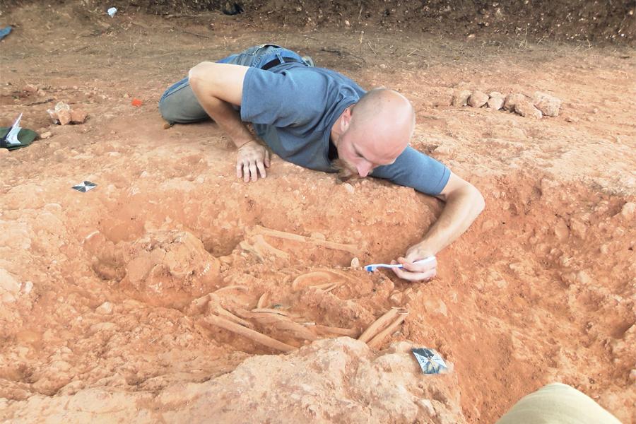 A student lays in the dirt over a skeleton that he's uncovering with a toothbrush.