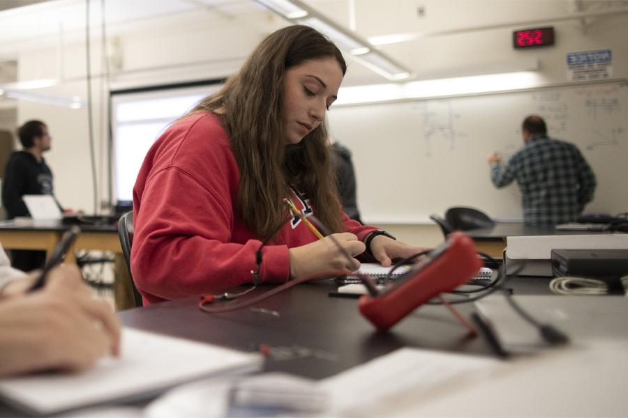 A student in a red shirt sits at a work table and takes notes on a notepad.