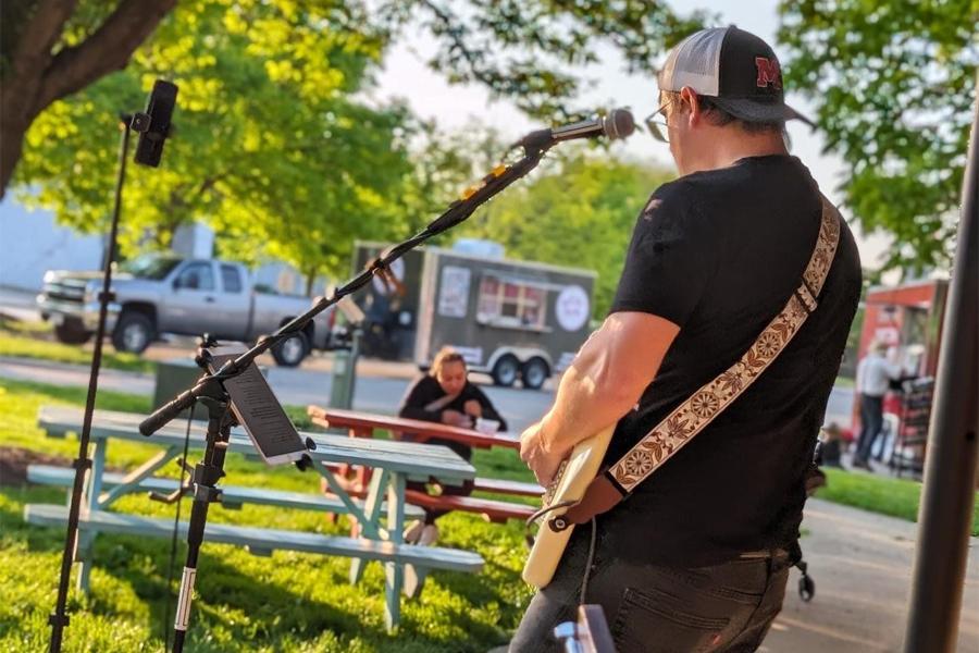 A person plays a guitar outdoors for a small crowd with a food truck in the background.