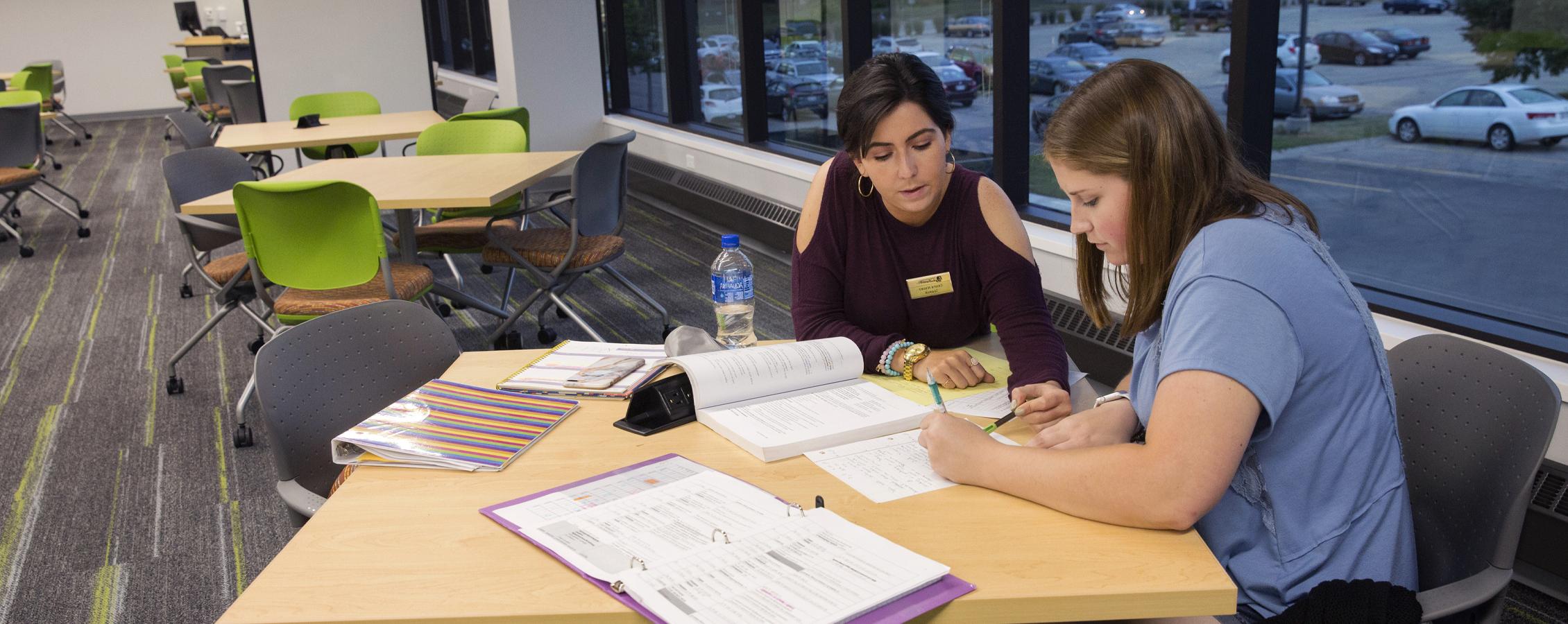 An international student from Venezuela tutors another student at a table.