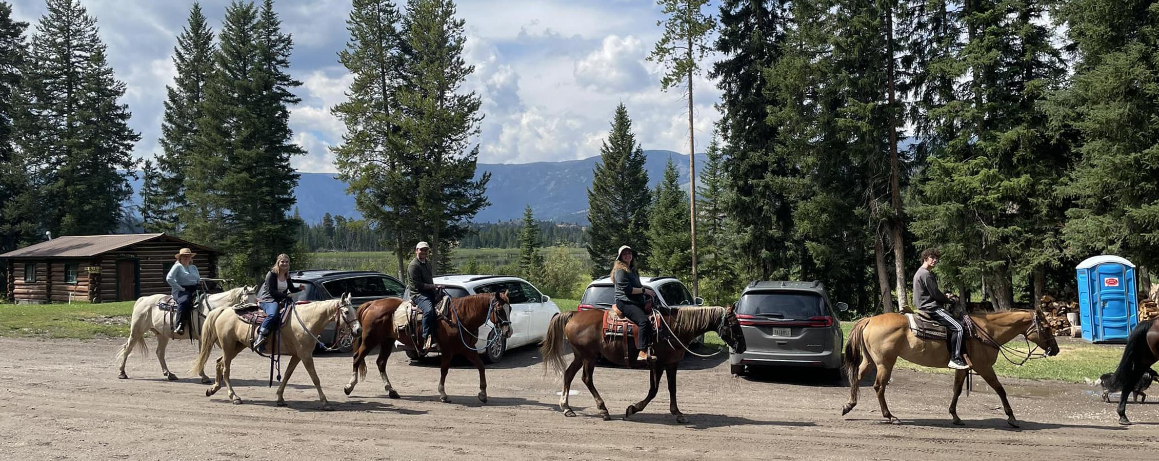 A group of people ride horses amidst mountains and evergreen trees.